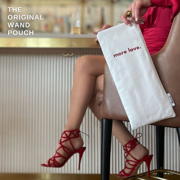 Woman seated at a bar holding the Quiver.love “More Love” wand pouch in white reclaimed denim with bold red glitter text; stylish and discreet storage for wellness essentials adn adult toys